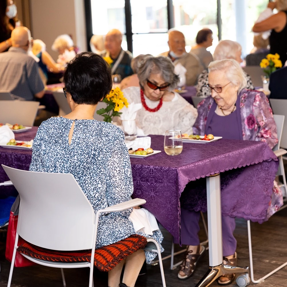 Some residents enjoying a healthy meal for seniors together to celebrate Wisteria Place. They are in fancy attire and there is a nice purple tablecloth on the tables.
