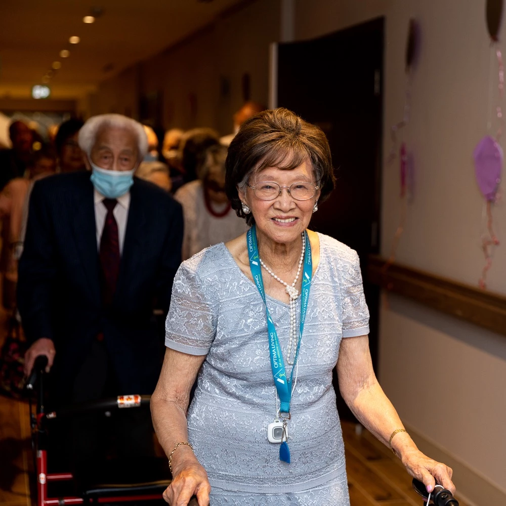 Senior Woman walking down the hallway smiling of a senior home. She has an Optima Living lanyard on and has other residents walking behind her.